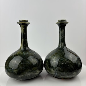 Pode incluir: Dois vasos de cerâmica verde escuro com um padrão texturizado semelhante a uma rede. Os vasos têm um corpo arredondado e um pescoço longo e fino com um topo alargado. Os vasos são exibidos em uma superfície branca.