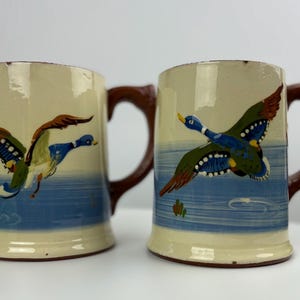 May include: Two ceramic mugs with brown handles and blue and white painted designs of ducks flying over a blue lake.