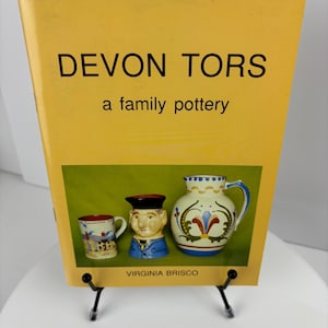 Könnte beinhalten: Ein gelbes Buch mit dem Titel "Devon Tors, eine Familien-Töpferei" von Virginia Brisco. Der Umschlag zeigt ein Foto von drei Töpferwaren: einer Tasse, einer Kanne und einer Kopf-Tasse.