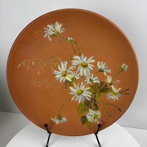 Può includere: Un piatto in terracotta con un disegno dipinto a mano di margherite bianche e foglie verdi. Il piatto è su un supporto in metallo nero.