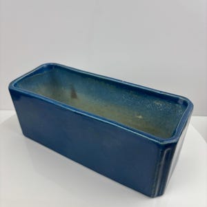 Puede incluir: Una jardinera rectangular de cerámica en un tono azul oscuro. La jardinera tiene una superficie ligeramente texturizada y una forma rectangular. El interior tiene un color azul más claro y descolorido. Adecuado para uso en interiores o exteriores.