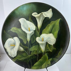 Könnte beinhalten: Eine runde, dunkelgrüne dekorative Platte mit einem Gemälde von weißen Calla-Lilien mit gelben Zentren und grünen Blättern. Die Platte wird auf einem schwarzen Metallständer präsentiert, der das florale Kunstwerk zeigt.