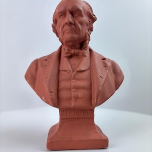 May include: A terracotta bust of a man in a suit and bow tie. The bust is mounted on a rectangular base with the name "GLADSTONE" inscribed on the front. The sculpture is a reddish-brown colour.
