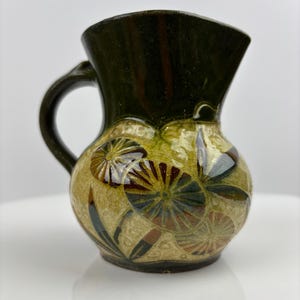 Puede incluir: Jarra de cerámica pequeña, hecha a mano, con la parte superior y el asa de color verde oscuro. El cuerpo de la jarra es de color beige con un diseño floral en tonos verdes, marrones y azules. La jarra tiene una base redondeada y un pico acampanado.