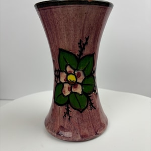 Peut inclure: Un petit vase en céramique rose avec un motif floral. Le vase a un col étroit et une base large. La fleur est blanche avec un centre jaune et des feuilles vertes.