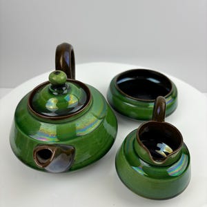 Puede incluir: Un juego de tetera, jarra de crema y azucarero de cerámica verde con glaseado iridiscente y detalles marrones.