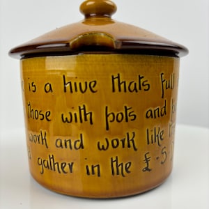 May include: A brown ceramic sugar bowl with a lid. The bowl has a brown and gold glaze and a quote written on the side: "is a hive thats full of those with pots and pans, work and work like the bees and gather in the £.5"