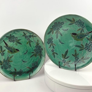 May include: Two green ceramic plates with bird and floral designs. Each plate features a central bird image surrounded by a wreath of flowers and leaves. The plates are displayed on black metal stands.