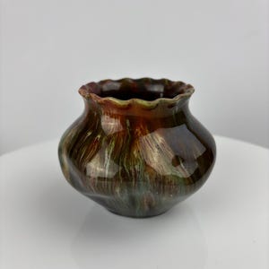 Pode incluir: Um pequeno vaso de cerâmica marrom, verde e dourado com uma borda festonada. O vaso tem um esmalte marmorizado.