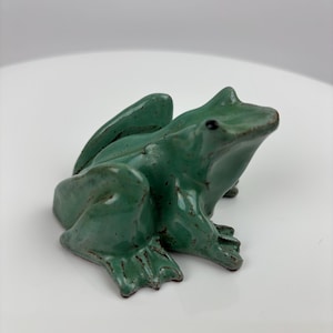 May include: A ceramic frog figurine in a sitting position. The frog is a mottled green color with dark brown accents. The figurine has detailed features, including webbed feet and bulging eyes. The frog is sitting on a white surface.