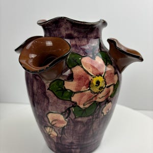 Könnte beinhalten: Eine dekorative Keramikvase mit dunkelvioletter Basis und gerüschtem Rand. Die Vase hat ein handgemaltes Blumendesign mit rosa und gelben Blüten und grünen Blättern. Sie hat drei braune, blütenblattförmige Öffnungen.