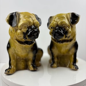 Puede incluir: Dos figuras de perro pug de cerámica. Los pugs están sentados erguidos con una combinación de colores marrón y negro. Los perros tienen narices y orejas negras. Las figuras están sobre una superficie blanca.