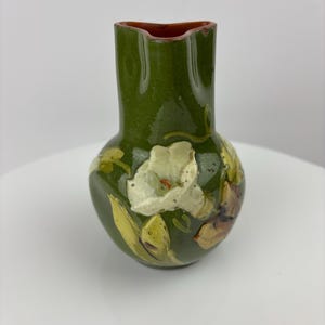 May include: A small, green ceramic vase with a bulbous base and a narrow neck. The vase features a raised floral design with white and yellow flowers and brown accents. The interior rim is a reddish-brown color.