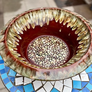 May include: A large, round, brown ceramic bowl with a green and brown speckled glaze. The bowl has a wide rim and a deep interior. The bowl is sitting on a mosaic tile surface.