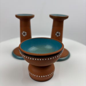 Puede incluir: Un juego de tres piezas de cerámica de color terracota. El juego incluye un cuenco pequeño con interior turquesa y dos candelabros a juego. El cuenco y los candelabros tienen diseños de puntos y estrellas blancos.