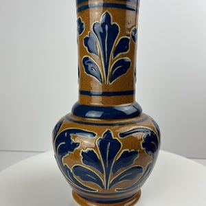 Puede incluir: Un jarrón de cerámica marrón con un diseño floral azul. El jarrón tiene una base ancha y un cuello estrecho.