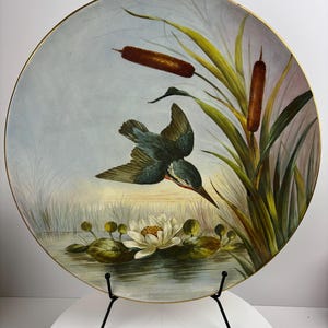 May include: A round ceramic plate with a hand-painted design of a blue bird flying over a white water lily in a pond with green reeds and brown cattails.