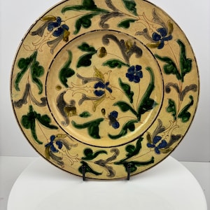 Op de afbeelding: Een decoratieve keramische schaal met een crèmekleurige achtergrond en ingewikkelde bloempatronen. Het ontwerp bevat groene bladeren, blauwe bloemen en grijze accenten. De schaal wordt gepresenteerd op een zwarte metalen standaard.
