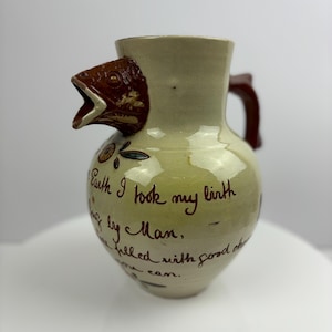 May include: A ceramic pitcher with a bird head spout and a brown handle. The pitcher is decorated with brown and green floral designs and the inscription "Earth I took my birth by Man, filled with good cheer I am."