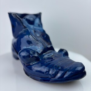 May include: A dark blue ceramic boot sculpture with a glossy finish. The boot features a rounded toe, a small heel, and a folded-over top. The surface reflects light, emphasizing the curves and details of the boot.