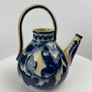 Peut inclure: Une théière en céramique avec un corps arrondi, une anse incurvée et un bec verseur. La théière présente une base de couleur crème avec des motifs floraux et des accents bleu foncé. La anse et le bec sont également crème avec des détails bleus.