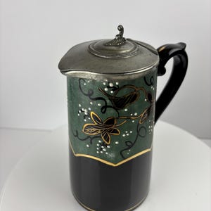 May include: A green and black ceramic pitcher with a pewter lid. The pitcher has a floral design with gold accents.