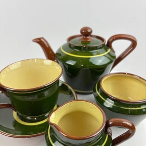 May include: A green and brown ceramic tea set comprising a teapot, teacup and saucer, creamer, and sugar bowl. The set features a yellow stripe and interior. The teapot has a brown handle and lid. The teacup has a matching saucer.