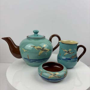 Op de afbeelding: Een tealblauwe theepot, roomkan en suikerpot met bruine handvatten en een zeemeeuwendesign. De theepot heeft een bruin deksel en de suikerpot heeft een bruine binnenkant.