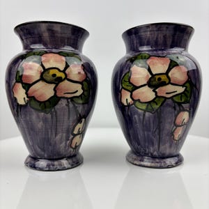 Könnte beinhalten: Zwei kleine, violette Keramikvasen mit Blumendesigns. Jede Vase zeigt eine rosa Blume mit grünen Blättern und einer gelben Mitte. Die Vasen haben eine strukturierte, gestreifte Oberfläche.