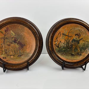 May include: A pair of antique decorative plates in wooden frames. One plate shows a child with a basket near a tree, the other depicts a boy with a stick. Both plates have a warm colour palette and are displayed on black stands.