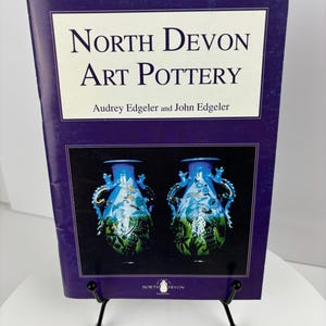 May include: A purple book titled "North Devon Art Pottery" by Audrey Edgelar and John Edgelar. The cover features a photograph of two blue and green ceramic vases with dragon designs.