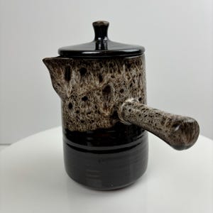 Puede incluir: Una cafetera de cerámica marrón y negra con tapa y asa. La cafetera tiene un esmalte moteado.