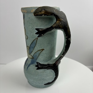 Puede incluir: Un jarrón de cerámica con un esmalte azul claro moteado y un asa única con forma de foca marrón oscuro. El jarrón presenta diseños florales pintados en azul y crema. El borde tiene un aspecto envejecido y desgastado.