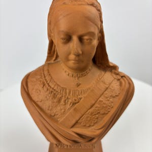 May include: A terracotta bust of Queen Victoria, featuring a detailed depiction of her face, hair, and attire. The bust includes the inscription "VICTORIA R.I." above the dates "1837-1897" and the phrase "GOD SAVE THE QUEEN".