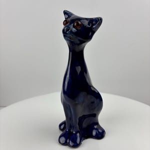 May include: A glossy, dark blue ceramic cat figurine with red eyes. The cat is in a seated position with its head tilted upwards. The figurine has a smooth, reflective surface and is a decorative item.