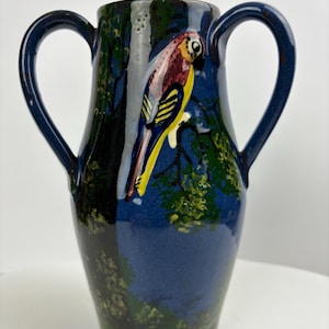 May include: A blue ceramic vase with two handles and a painted parrot design. The parrot is yellow, red, and black with a blue background.