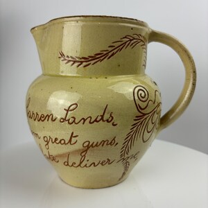 Puede incluir: Una jarra de cerámica amarilla con asa marrón y diseños pintados en marrón. La jarra tiene una cita que dice "Barren Lands, from great guns, we deliver fire".