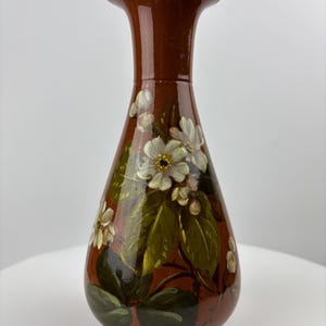 May include: A brown ceramic vase with a flared top and a rounded body. The vase is hand-painted with white flowers, green leaves, and small white buds. The vase is approximately 25 cm tall.
