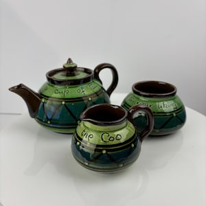 Op de afbeelding: Een groene en bruine keramische theeset met een theepot, roomkan en suikerpot. De theepot heeft de tekst "a cup of tea" erop, de roomkan heeft de tekst "the Coo" erop en de suikerpot heeft de tekst "to your liking" erop.