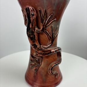 May include: A red and brown ceramic vase with a lizard design. The lizard is sculpted in relief and has a black and brown glaze.
