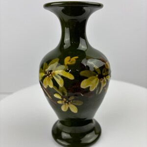 May include: A dark green ceramic vase with a glossy finish. The vase features hand-painted yellow and white floral designs. The vase has a flared top, a rounded body, and a circular base. The vase is approximately 18 cm tall.