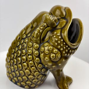 May include: A ceramic frog-shaped wine bottle holder in a glossy olive green glaze. The frog has a textured, bumpy skin pattern and an open mouth to hold a bottle. The holder is designed with a footed base and detailed features.