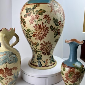 May include: Three ceramic vases with floral designs. The largest vase is tall with a wide base, featuring a cream background with red, brown, and green flowers. The other two vases are smaller, with similar floral patterns and a blue top.
