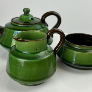 Puede incluir: Un juego de tetera, jarra de crema y azucarero de cerámica verde. La tetera tiene una tapa y un asa marrones. La jarra de crema y el azucarero tienen asas marrones.