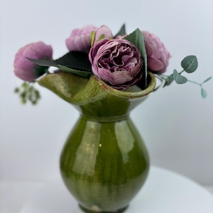 May include: A green ceramic vase with a glossy finish, holding a bouquet of pink and purple artificial flowers. The vase has a unique, flared top and a rounded body, creating a decorative floral arrangement.