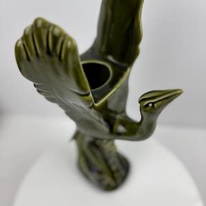 Può includere: Una statuetta di uccello in ceramica con finitura verde oliva lucida. L'uccello è raffigurato con le ali spiegate e un'apertura circolare è visibile al centro. La statuetta è appoggiata su una superficie bianca.