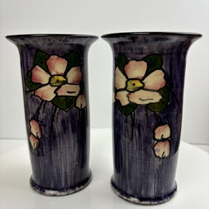 Peut inclure: Deux vases en céramique violette avec des motifs floraux. Chaque vase présente une fleur rose avec un centre jaune et des feuilles vertes.