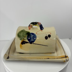 Könnte beinhalten: Eine Butterdose aus Keramik mit einem Deckel mit Griff und einem abstrakten, bemalten Design in Blautönen, Grün, Gelb, Orange und Schwarz. Die Schale steht auf einem passenden quadratischen Teller.