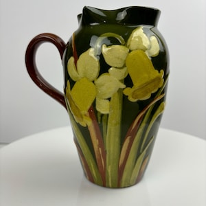 May include: A green ceramic jug with a brown handle and a floral design of yellow daffodils.