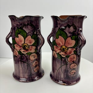 Peut inclure: Deux vases en céramique vintage avec des motifs floraux. Les vases sont violets avec des fleurs roses et vertes et ont deux poignées de chaque côté.